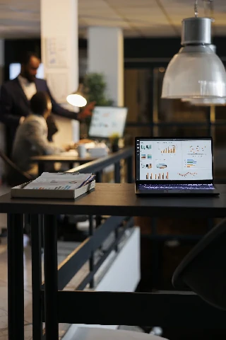 laptop computer with financial graphs screen standing table startup office coworkers discussing company strategy background employees working late night startup office1